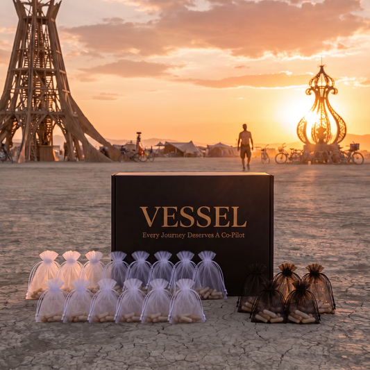 A black Vessel gift box with gold lettering stands on a cracked desert ground at sunset, with small sheer gift bags filled with nuts arranged in front. The background shows a large wooden structure and a person walking in the distance under a warm orange sky.